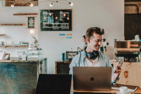 A digital marketer smiling while working on a MacBook, looking at his phone, reflecting on his digital footprint and online presence.