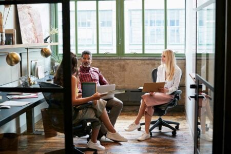 A Digital Marketing Manager and two colleagues are sitting together in an office meeting room, engaged in a discussion about a project. They are seated around a table with laptops and documents in front of them, collaborating as a team.
